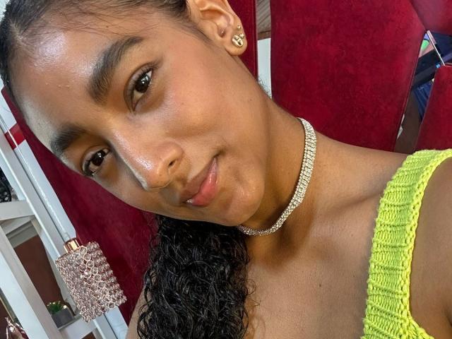 Close-up selfie of NatalyBlonder with dark skin, curly black hair, and a shiny yellow top. She wears a diamond choker, smiling subtly, with visible sweat on her face. Red background.