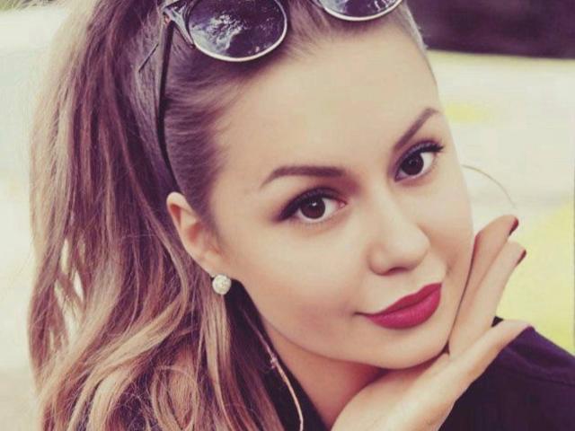 Close-up of CrystallyEyes with her hand touching her cheek, brown hair in a ponytail, sunglasses on top, red lipstick, dark eyeliner, pearl earrings, smiling, slightly blurred background. Fucking sexy.