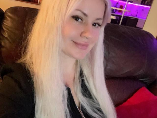 Close-up van CrystallyEyes, een blonde vrouw met lang, steil haar, die lichtjes glimlacht en een zwarte top draagt, zittend op een donkere leren bank met een rode kussen. Neonpaarse lichten op de achtergrond.