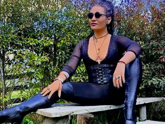 DivaMarie, assise sur un banc en bois, portant un haut noir transparent, un pantalon en cuir noir moulant, des bottes, et des lunettes de soleil, de longs cheveux noirs, et de multiples colliers, dégageant une aura de confiance et de sensualité..