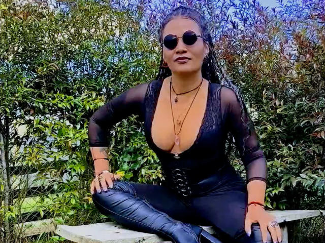 DivaMarie, wet hair, wearing black sheer top with deep cleavage, black pants, sitting cross-legged on a bench, sunglasses, necklace, outdoor, green foliage background, confident, sexy, casual pose.