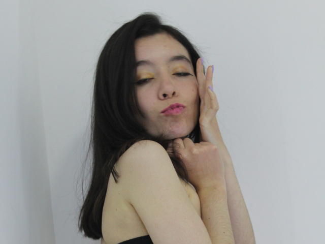 ElodiePerrine has long black hair, pale skin, and yellow eyeshadow. She's touching her face with one hand, lips slightly parted. She's wearing a black top, eyes half-closed. Fucking sexy.