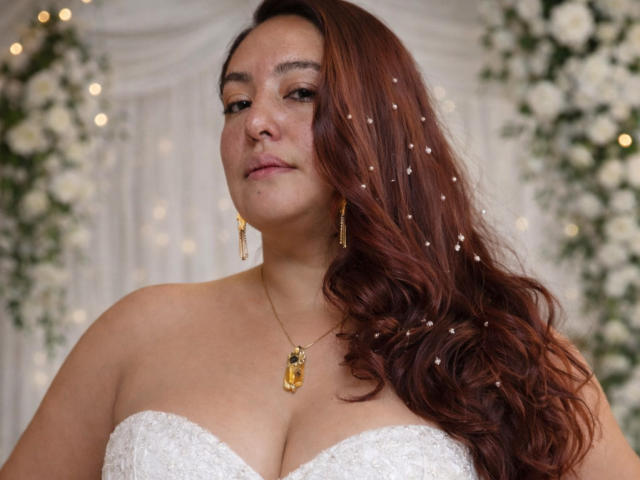 TerraMegaBoobs, formosa con grandi seni, indossa un abito bianco senza spalline in pizzo e ha lunghi capelli castani ondulati e scintillanti. perline, orecchini d'oro e collana. Lo sfondo presenta fiori bianchi sfocati e luci soffuse..