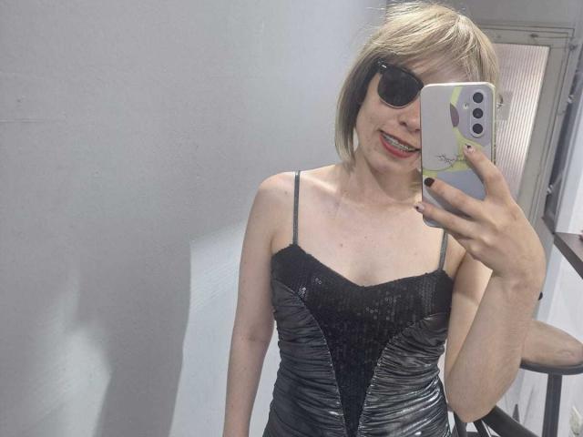 ElodiePerrine, short blonde bob, black sunglasses, red lipstick, black sequin dress, holding iPhone with white case, mirror selfie, sexy, confident, visible cock outline in dress, bright indoor lighting.