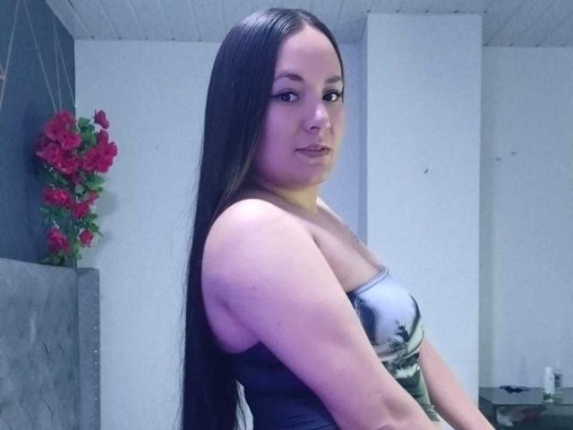 Foto van AryNoa met lang zwart haar, die een glanzende zilveren top zonder bandjes draagt en die over haar schouder kijkt. een lichte glimlach.
rode rozen op de achtergrond. Blauwe verlichting.