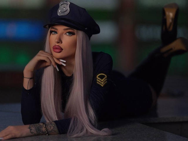 JuicyJessye, con cabello largo, lacio y lavanda, vestida con un gorro y uniforme de policía negros, acostada sobre una mesa, con las piernas levantadas y una mano... cerca de la boca, labios rojos, tatuaje en el antebrazo, aspecto sexy, mirada seductora..