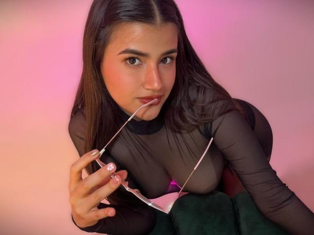 Fotografia di Iris Blair, una giovane donna con lunghi capelli neri, che indossa una maglia nera trasparente e sta succhiando un oggetto di vetro trasparente. L'illuminazione rosa e arancione crea un'atmosfera sensuale. Il suo seno è visibile attraverso il tessuto..