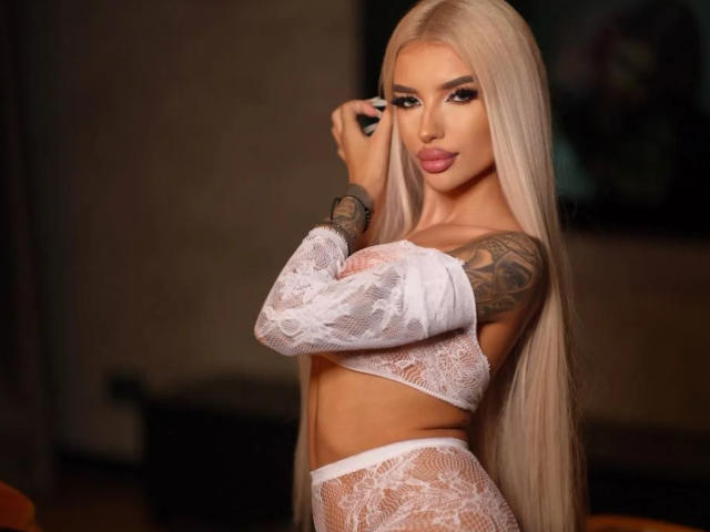 JuicyJessye with long, straight platinum blonde hair, wearing white lace lingerie, has a tattoo on her right arm, posing with one hand on her face, sultry expression, dim lighting.