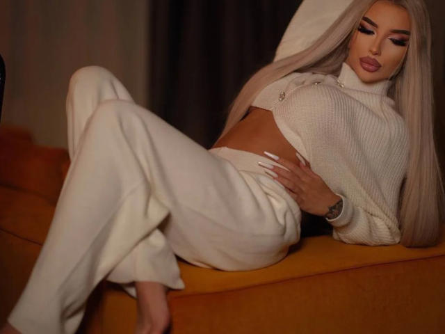 JuicyJessye est allongée sur un canapé orange, vêtue d'un pantalon blanc et d'un pull blanc court. De longs cheveux blonds platine, des lèvres pulpeuses et des ongles soignés. Elle expose sa taille et ses jambes. Ambiance sexy et pleine d'assurance..