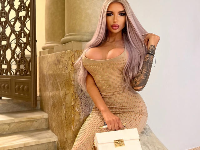 JuicyJessye with long, platinum blonde hair, wearing a tight, beige, mesh dress, showcasing large breasts and curvy figure, leaning against a stone wall, holding a white purse, sexy pose.