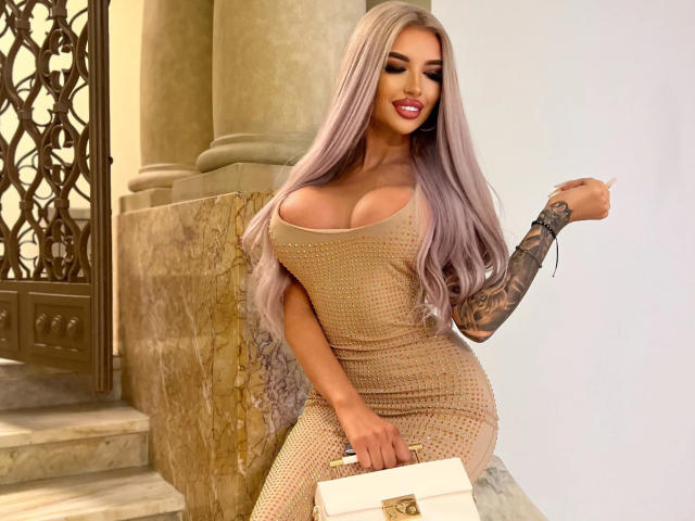 JuicyJessye, avec ses longs cheveux lisses lavande, porte une robe beige moulante en maille qui met en valeur sa poitrine généreuse. Elle se penche contre un mur de pierre, la main levée, son bras tatoué visible, dans une pose séduisante..