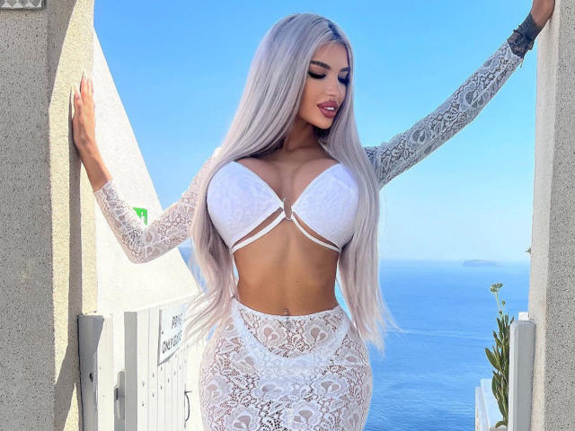 JuicyJessye, avec ses longs cheveux argentés, ses grandes seins dans un soutien-gorge en dentelle blanche, et une jupe assortie en dentelle, debout, les bras s'étendant entre des piliers blancs, avec l'océan et un ciel bleu vif en arrière-plan..