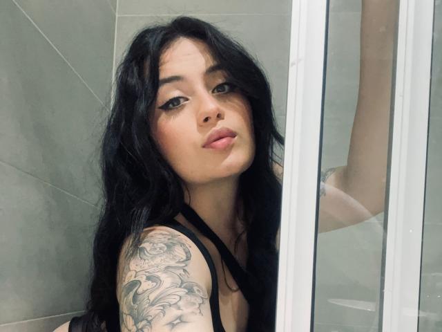 AnnyBentleii, a young woman with long black hair and tattoos on her right arm, leans against a glass wall with bright vertical lights, looking directly at the camera with a sultry expression.