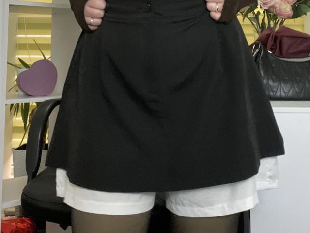 Foto em close-up de Madrinia vestindo uma saia preta com shorts brancos por baixo, com as mãos segurando a saia. Cadeira preta, objeto roxo em formato de coração, bolsa preta e flores rosa ao fundo. Muito atraente..