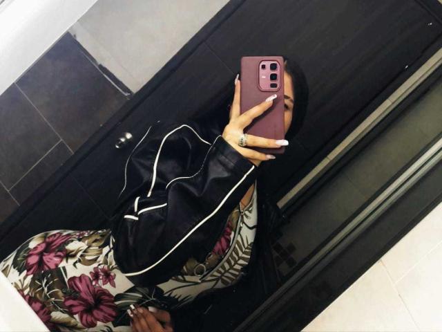 Alana Mathews tira uma selfie em um espelho, segurando um telefone rosa com uma capa preta. Ela está vestindo uma blusa com estampas florais e um casaco preto, com fones de ouvido brancos visíveis. Muito quente..