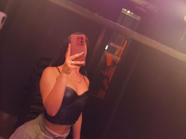 Alana Mathews che si fa un selfie in una stanza poco illuminata, tenendo un telefono rosso con una mano, indossando un top nero senza spalline e pantaloni bianchi.
scollatura visibile, illuminazione rossa, posa informale.