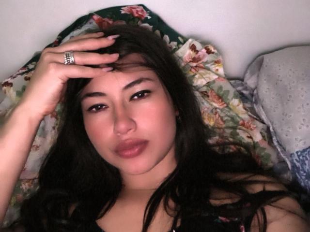 Foto van Alana Mathews, liggend op een bed met bloemenkussens, hand op haar voorhoofd, donker haar, bruine ogen, licht open. lippen, draagt een top met bloemenprint, ring aan de vinger, sexy, ontspannen.