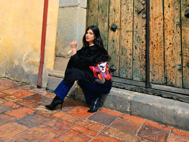 Paulina Valley se apoya contra una puerta de madera desgastada, vistiendo un abrigo negro y botas, y lleva un bolso de mano con un estampado rojo y blanco. Pared de color amarillo brillante y pavimento de ladrillo rojo. Pose increíblemente atractiva..