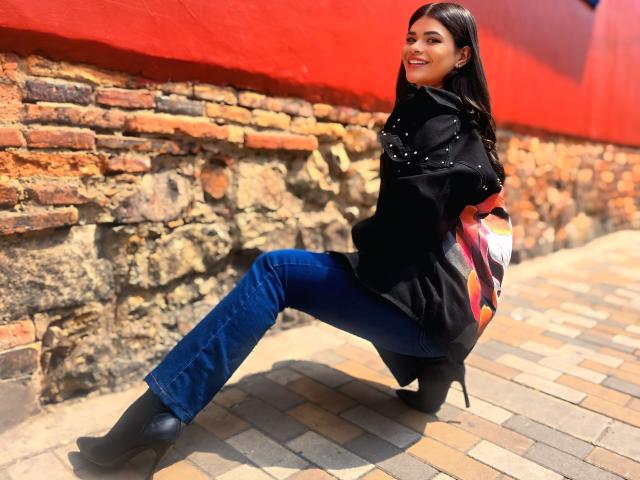 PaulinaValley lehnt sich an eine Ziegelwand, vor einem hellroten Hintergrund, und trägt einen schwarzen Mantel, blaue Jeans und schwarze Stiefel. Sie lächelt, mit einem Bein angehoben und den Händen hinter dem Rücken. Sonnenlicht betont ihre dunklen Haare..