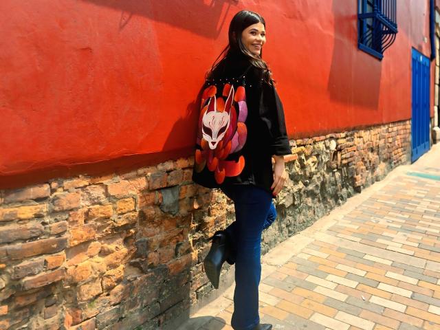 PaulinaValley with long dark hair, black jacket, and red backpack with white cat design, leans against a bright red wall and stone base. Sunlight casts sharp shadows, street path visible.