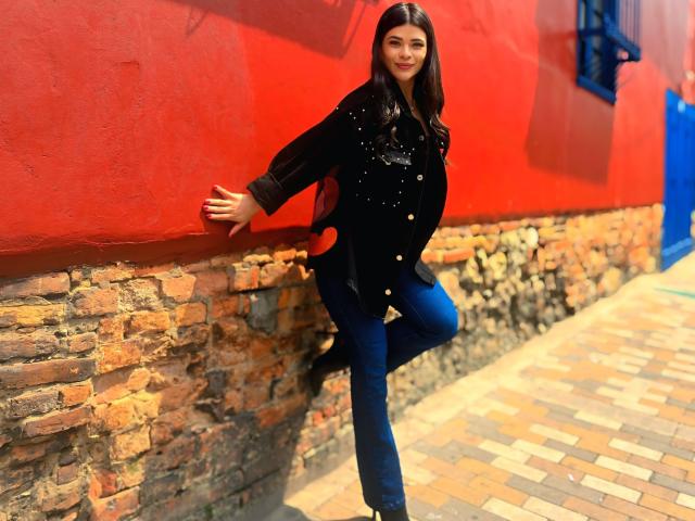 PaulinaValley, com cabelo escuro e um casaco preto de botões, está encostada em uma parede vermelha, com uma perna levantada. Ela está vestindo calças jeans azuis e está em pé em uma rua pavimentada com tijolos. Pose incrivelmente sexy..