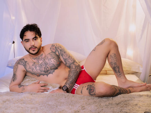 Dante Dimarco, sem camisa, com tatuagens cobrindo o peito e os braços, deitado em uma cama branca, usando roupa íntima vermelha, com o pênis visível. através do tecido, olhar intenso, cabelo cacheado escuro, relógio no pulso, iluminação suave.