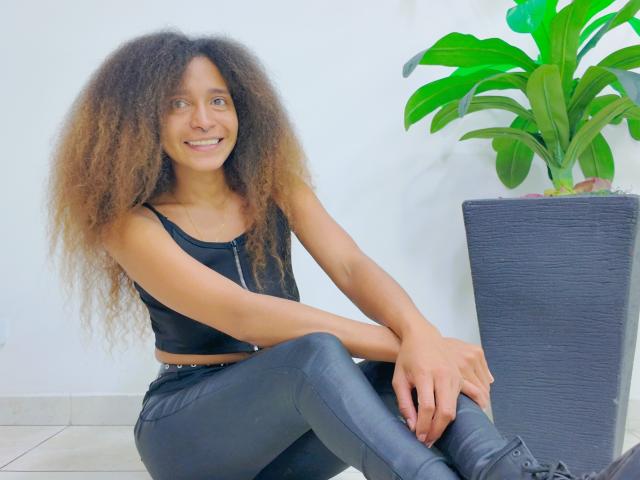 Fotografía de una atractiva SweetSexx con cabello castaño largo y rizado, vestida con una camiseta negra y jeans azules ajustados, sentada piernas cruzadas, apoyada en una maceta negra con una planta verde..