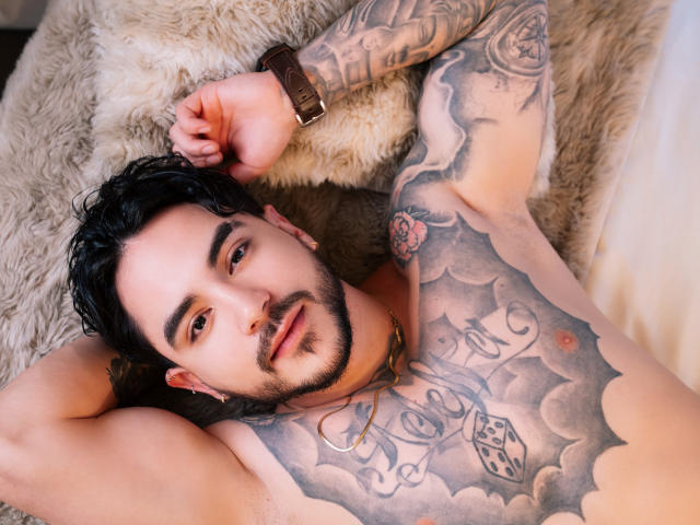 DanteDimarco, met tatoeages, ligt op een pluizige deken, zonder shirt, met zwart haar en baard, en kijkt naar de camera. Gedetailleerde borsttatoeages, inclusief wolken en dobbelstenen. Hand op het hoofd, horloge om de pols..