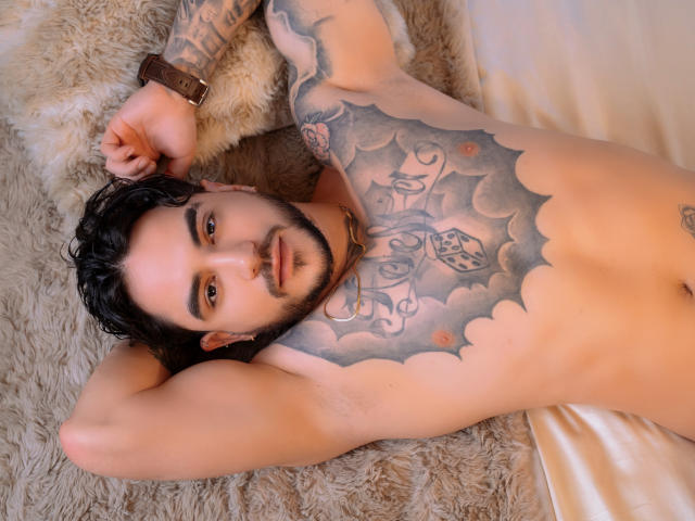 DanteDimarco lies on a furry rug, shirtless, with a large cloud and bird tattoo covering his chest. He's looking up, hand behind head, brown hair, beard, muscular, circumcised, erect.