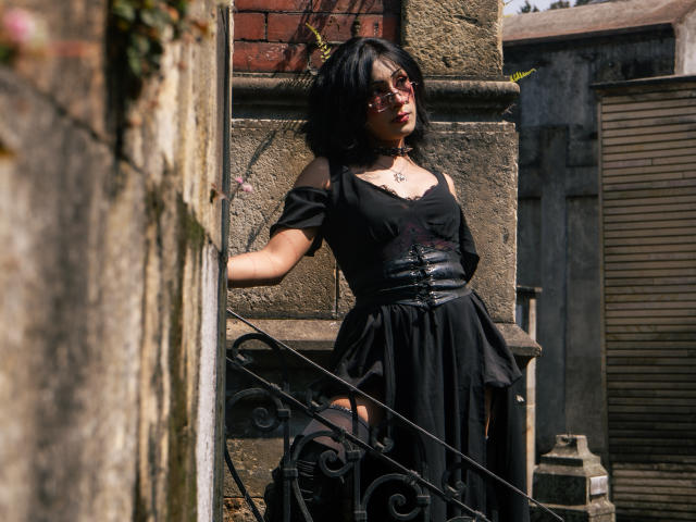 NoemiSky with black bob haircut, wearing off-shoulder black dress and black corset, leaning against stone wall, sunlight highlights her figure, shadows add depth, urban alley setting, slight sexual tension.