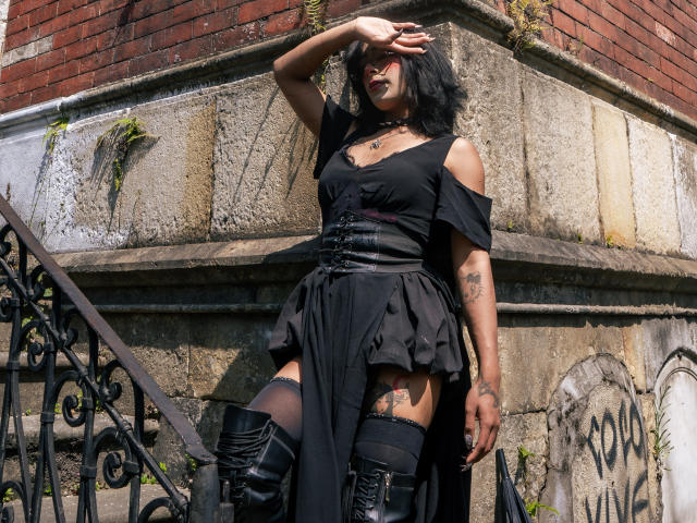 NoemiSky, mit dunklem Haar, steht vor einer verwitterten Steinmauer und schützt ihre Augen vor dem Licht. Sie trägt ein schwarzes Oberteil mit einem Schulterauschnitt, einen gerüschten Rock, hohe Stiefel und hat Tattoos. Das Sonnenlicht betont ihre selbstbewusste, sinnliche Pose..
