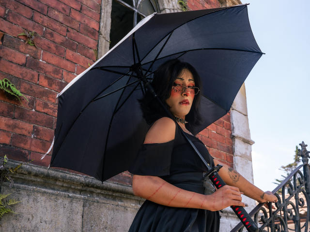 NoemiSky tient un parapluie noir devant un mur de briques rouges, vêtue d'une robe noire à une épaule, avec du rouge à lèvres et un maquillage subtil. lunettes. Son bras droit porte un tatouage visible. Une ambiance vraiment intense..