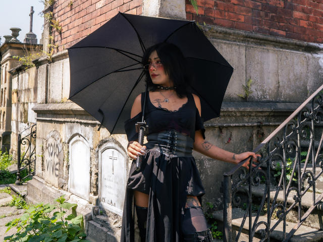 NoemiSky staat daar, een zwarte paraplu in haar hand, gekleed in een zwarte corset en rok, met zichtbare tatoeages. Ze staat naast een bakstenen muur, en het zonlicht verlicht haar. Ze kijkt naar de zijkant..