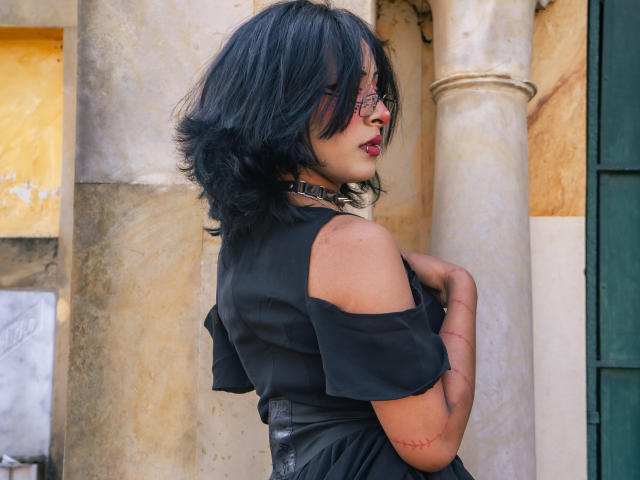 NoemiSky, a woman with black shoulder-length hair, red lipstick, and glasses, wearing a black off-shoulder top, stands against a beige column, touching her chest with a sexy, sultry expression.