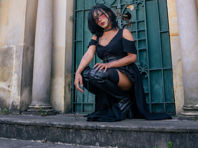 NoemiSky squats on stone steps, wearing a black off-shoulder dress and thigh-high boots. She has dark hair, red lipstick, and black choker. Background: aged green door and columns. Sexy, intense vibe.