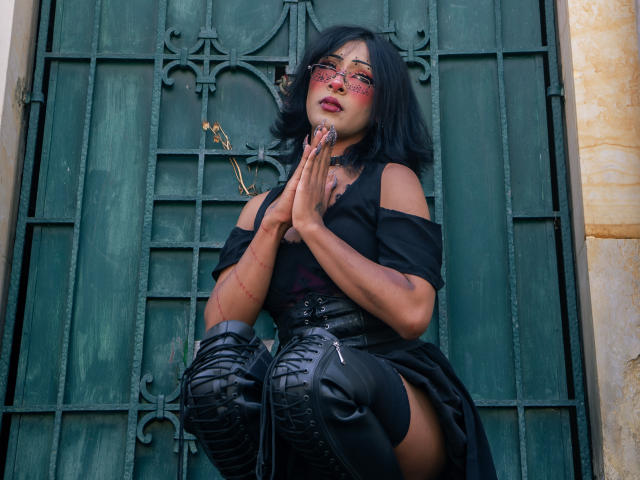 NoemiSky, una mujer de pelo oscuro con gafas rojas, se arrodilla frente a una puerta de metal verde, con las manos juntas en señal de oración. posición. Ella lleva un vestido negro con un hombro descubierto y botas que le llegan hasta el muslo. Sus labios están pintados de rojo..