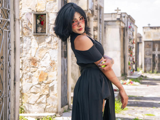 NoemiSky, une femme séduisante avec des cheveux noirs et des lunettes rouges, se tient dans une ruelle étroite, vêtue d'une robe noire à une épaule. Elle a un tatouage sur son bras gauche, qui la regarde avec une expression séduisante..