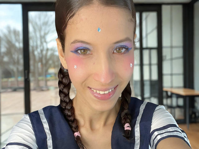 Close-up photo of TheDollZarita, a smiling woman with braided hair, purple eyeshadow, and pink star-shaped face glitter. She's wearing a black and white sailor uniform, cock visible in the background.
