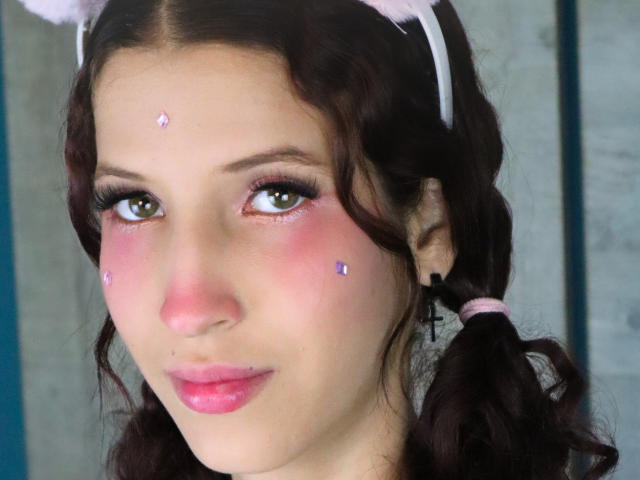 Gros plan de TheDollZarita, une jeune femme à la peau claire, avec des cheveux noirs et bouclés attachés en couettes, un blush rose et des étoiles violettes. des joues rosées. Elle a une expression séduisante, avec des lèvres roses et pulpeuses..