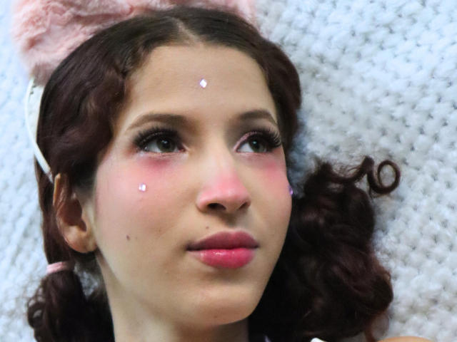 Close-up foto van TheDollZarita, een jonge vrouw met lichte huid, donker krullend haar en roze lippen, liggend op een wit, gestructureerd oppervlak. oppervlak. Glitterende accenten op haar voorhoofd, intense blik omhoog..