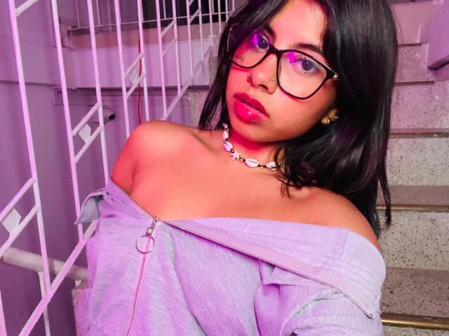 Photograph of CelesteMorel, with long black hair, wearing glasses, off-shoulder light gray top, necklace, and red lipstick. Purple light casts glow on face, leaning against white bars. Fucking sexy.