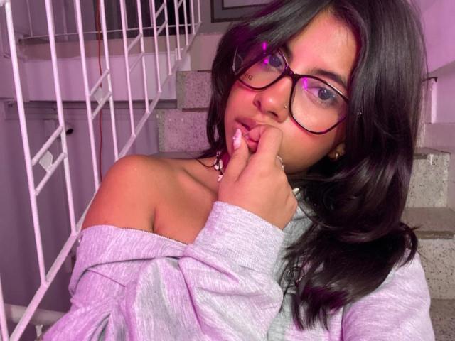 Photograph of CelesteMorel, a sexy woman with long black hair, wearing glasses and a white off-shoulder sweater, biting her finger seductively, with neon pink highlights in her hair and glasses.