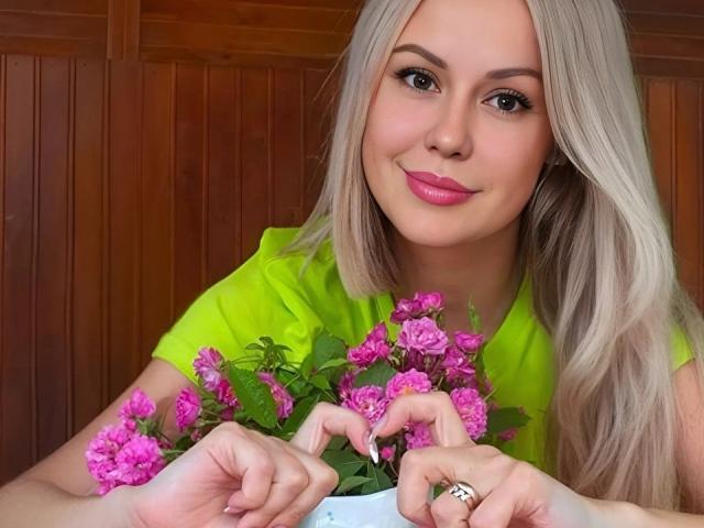 CrystallyEyes, blond, met roze anjers, heldergroen shirt, houten achtergrond, lichte glimlach, roze lippenstift, verzorgde nagels, ring aan de linkerhand, close-up, zacht licht, seksuele spanning, nonchalante pose, levendige kleuren.