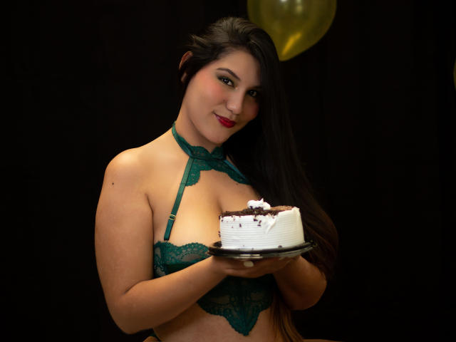 Maria Rowe, una mujer de cabello oscuro y piel clara, vestida con un sujetador verde de encaje, sosteniendo una rebanada de pastel blanco cubierto de glaseado. Ella sonríe levemente, fondo oscuro, globo dorado arriba. Tensión sexual, pose sugestiva..