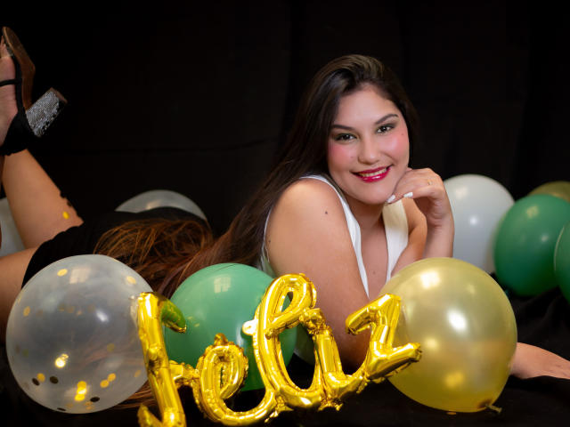 Maria Rowe, allongée sur le ventre, souriante, avec de longs cheveux noirs, portant un haut blanc, entourée de vert et de blanc, des ballons jaunes, et un panneau lumineux avec le chiffre "QUATRE" devant elle.