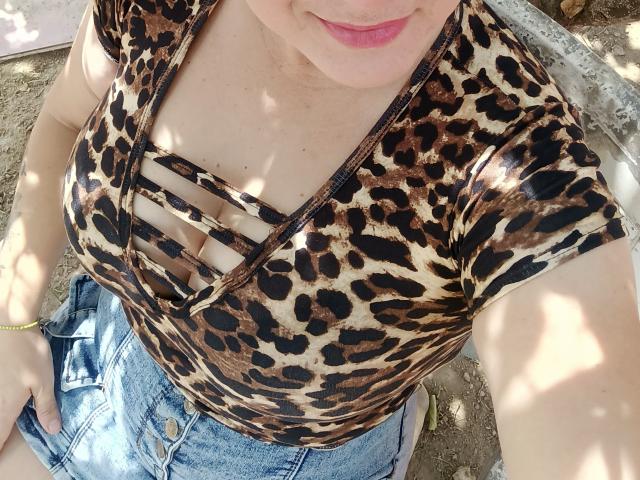 Close-up photo of AlexaHotSeins with a leopard print top, showing cleavage and cut-out design, wearing high-waisted jeans. Dappled sunlight on skin, pink lips slightly visible, casual outdoor setting.