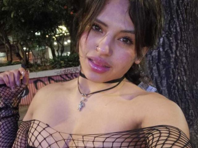 EmilieJoy, with brown hair and nose ring, wears fishnet top exposing small breasts, shiny pink lips, black choker with silver pendant, leaning against tree, nighttime urban setting, seductive gaze.