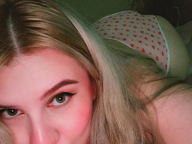 Close-up of Akkanee's face with intense brown eyes, partially obscured by long blonde hair. Wearing white panties with red heart patterns, lying on her side. Bright red cheek highlights. Sexual tension.