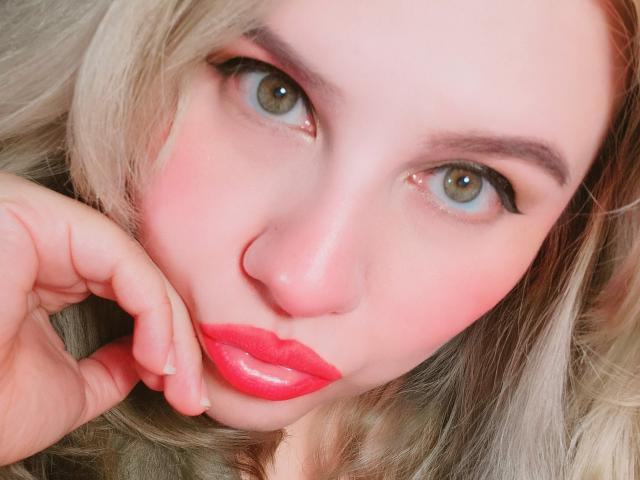 Close-up of Akkanee's face with light blue eyes, full red lips, and blonde hair. Akkanee's hand touches her chin, creating a seductive expression. Bright, intense colors. Sexual tension.
