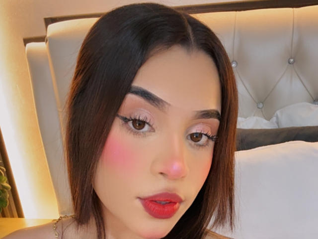 Close-up of LunaVali with fair skin, dark straight hair, bold red lips, pink blush, black eyeliner, and nude eyeshadow. She's sitting on a tufted beige headboard, looking directly at the camera. Her expression is seductive.